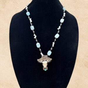 Blue Sky Longhorn necklace featuring large, pale blue Amazonite stones, lustrous freshwater pearls, and shimmering faceted crystal beads connected by silver-toned links. The statement pendant features a carved bone longhorn skull with ornate Tibetan silver detailing, three raised floral accents, and a turquoise gemstone. The reverse side features an intricate floral design.