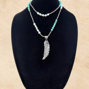 Feather and Pearls necklace featuring an intricately detailed silver-toned feather pendant with a turquoise stone accent and decorative detailing on the reverse. The necklace features vibrant turquoise beads, lustrous freshwater pearls, and shimmering crystal beads in a long, versatile strand that can be worn doubled for a layered look.